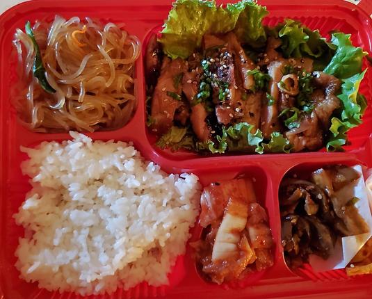 A red compartmentalized takeout container filled with various Korean dishes, including clear glass noodles, cooked white rice, sliced grilled chicken with sesame seeds and green onions over lettuce, kimchi, and braised mushrooms.