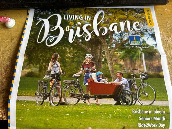 Photo of the front cover of the October 2025 issue of "Living in Brisbane" free magazine, showing a photo of several pushbikes and a cargobike carrying a small child in a grassy park with a bikeway and shady trees in the background, somewhere in the city.
