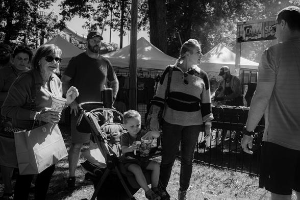 A black and white street photograph of a group of people at an outdoor festival or market. A young boy sits in a stroller in the center looking down at a travel cup while his family stands around him. To the left, an older woman in sunglasses looks past the camera, and to the right, another woman looks towards a vendor booth. In the background, there are more people, vendor tents under trees, and harsh sunlight casting strong shadows.