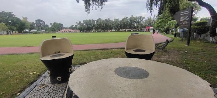 Outdoor park scene featuring a stone table and chair set, green lawn, trees, and a walking path.