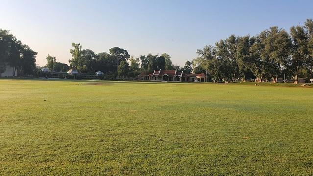 A wide green field with trees and buildings in the background.