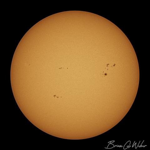 Full disk monochrome solar image colorized orange with assorted sunspots dotting the surface in several groups.