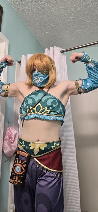 Me in my Gerudo Link cosplay! I've got a golden haired wig on and my lower chest/breast is covered over with a silky blue and gold garment. My forearms are covered up by the same type of fabric, and this extends to my biceps. Below the two belts, one green and one red with fringe, the Sheikah Slate hangs low because I didn't tighen my belt enough! I'm wearing a mask that conceals my lower face/jaw and there is a jewel dangling from my head onto my forehead.

In this pic, I am STRONK!! I'm doing the classic bicep flex with both arms raised. I love this one as it shows how much more slender I'm getting.