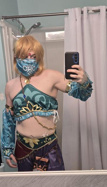 Me in my Gerudo Link cosplay! I've got a golden haired wig on and my lower chest/breast is covered over with a silky blue and gold garment. My forearms are covered up by the same type of fabric, and this extends to my biceps. Below the two belts, one green and one red with fringe, the Sheikah Slate hangs low because I didn't tighen my belt enough! I'm wearing a mask that conceals my lower face/jaw and there is a jewel dangling from my head onto my forehead.

In this pic, I have a fairly casual pose, holding my phone to the side while I take the pic through my mirror.