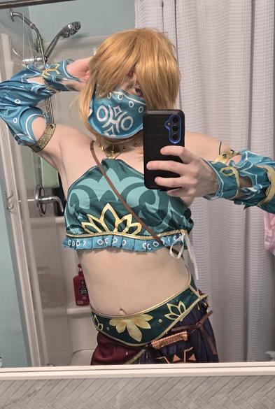 Me in my Gerudo Link cosplay! I've got a golden haired wig on and my lower chest/breast is covered over with a silky blue and gold garment. My forearms are covered up by the same type of fabric, and this extends to my biceps. Below the two belts, one green and one red with fringe, the Sheikah Slate hangs low because I didn't tighen my belt enough! I'm wearing a mask that conceals my lower face/jaw and there is a jewel dangling from my head onto my forehead.

In this pic, I'm taking a fabulous pose, with my left hand behind my head, as I stretch my torso backwards and to the right, while doing the opposite with my hips.