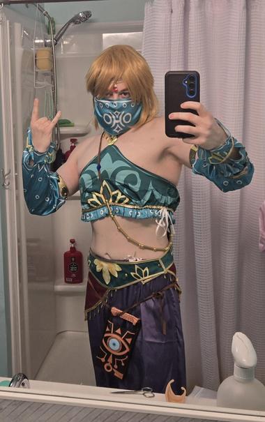 Me in my Gerudo Link cosplay! I've got a golden haired wig on and my lower chest/breast is covered over with a silky blue and gold garment. My forearms are covered up by the same type of fabric, and this extends to my biceps. Below the two belts, one green and one red with fringe, the Sheikah Slate hangs low because I didn't tighen my belt enough! I'm wearing a mask that conceals my lower face/jaw and there is a jewel dangling from my head onto my forehead.

I'm a metalhead as well, so I'm throwing horns in this pic!