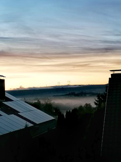 Das Foto zeigt eine ruhige Morgenstimmung mit Nebel über einer bewaldeten Landschaft. Im Vordergrund sind Hausdächer mit Solarpanels zu sehen, im Hintergrund erheben sich Windräder auf einem Hügel. Der Himmel ist in sanften Farben von Blau bis Gelb gefärbt – ein friedlicher Sonnenaufgang.