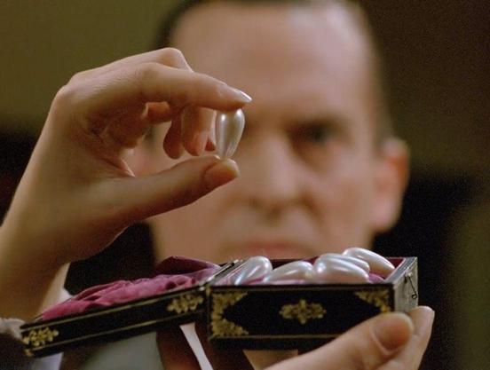 A woman's hand holding up a long pearl. There are more pearls in the ornate box in her hand. Behind the box, a stern-looking man watches