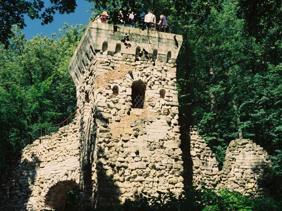 The image captures the ruins of a historical stone tower situated in a lush, green park. The structure stands tall with its weathered and eroded walls made from large stones, some sections missing entirely which expose brickwork underneath. Several arched openings are visible on different levels of the tower, suggesting former windows or passageways that have been worn down over time.

The topmost section features a flat platform where three individuals can be seen standing, surveying their surroundings. They seem minuscule in comparison to the towering ruins around them. To the both sides of the photo, there are additional structures appearing to be walls made from similar stone materials but appear even more weathered and less intact than the main tower.

The forest surrounding the tower is dense with green foliage, providing a stark contrast between nature reclaiming human-made structures and untouched natural beauty.