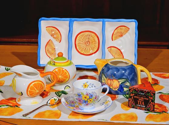 Invigorating tea tableau in bright orange and yellow colors with medium blue accents. There are a ceramic tray with citrus slices painted on it, vintage china, an orange themed tea towel, a cream and sugar set with designs of citrus fruits, a gold spoon with oranges painted on it. The tea is Sweet Orange Spice black tea.