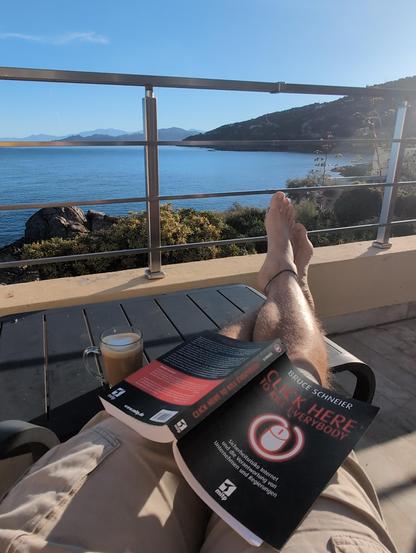 Blick vom Balkon auf Mittelmeer und Küstenlinie. Auf dem Schoß das Buch "Click here to kill everybody" von Bruce Schneider.