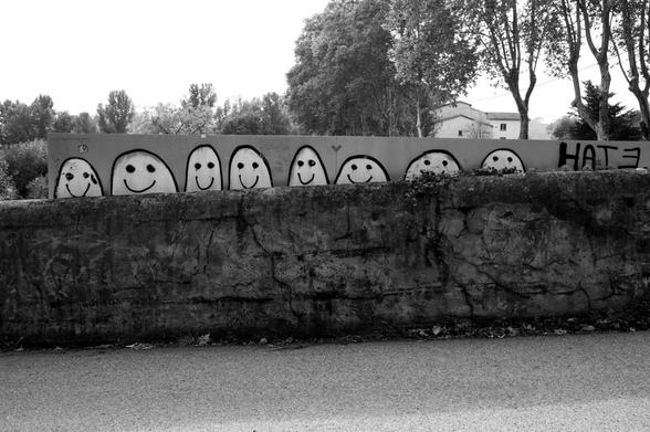 Bunch of little people and the word 'hate' graffitied behind a long sign above a low wall. (Version françæ) Bande de petiz bænæmmes et læ mot ‘hate’ (‘haine’) graffæs derrière æn lonx panneau al-dessus d’æn muret.
