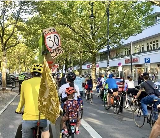 Auf der Fahrbahn des Kurfürstendamms in Berlin fährt eine Gruppe Radfahrende mit Kindern, anlässlich der KidicalMass am 20.09.2025. Ein selbst gebasteltes Schild fordert „City Speed Limit 30“.