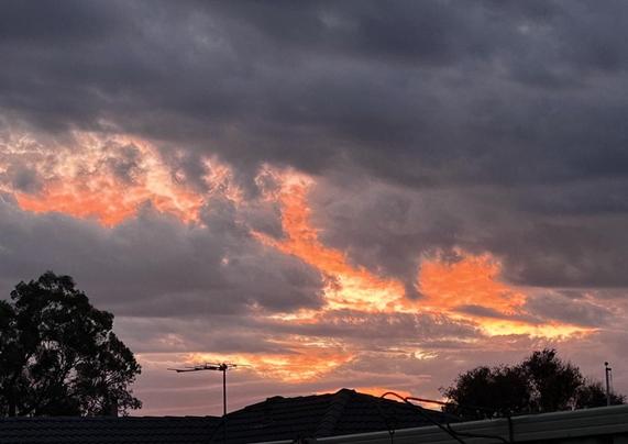Sunset tonight, dark storm clouds are in front on an amazing orange & yellow sunset. Trees & a roofline is framed against the sky.