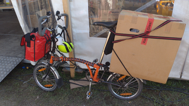 Deutschsprachige Bildbeschreibung:

Ein orange-schwarzes Brompton Faltraf mit einer roten Kurier-Umhängetasche auf dem vorderen Fahrer-Berlin-Front-Gepäckträger, einem neongelben Fahrradhelm am Lenker baumelnd und einem großen, nahezu quadratischen Karton auf dem hinteren Gepäckträger. Der Karton hat hinten ca. ⅓ Überhang und seine Oberkante ist in etwa 10cm oberhalb der Sitzfläche des Fahrradsattels. Es wird seitlich gehalten von den originalen Gepäckträger-Spannbändern, die schräg zu den Sattelschienen hoch führen. Waagrecht hält ein Spanngurt mit Ratsche das Paket an der Sattelstange fest.

English description:

An orange-black Brompton folding bicycle with a red messenger bag on its Fahrer-Berlin front rack, a yellow bicycle helmet hanging on its handlebar, and a huge carton box on rear rack. The box has about ⅓ overhang on the back and its top is about 10cm higher than the saddle. It's held by the Brompton's rack straps (connected to the saddle rails) and a ratchet strap around the whole box horizontally and the saddle post.