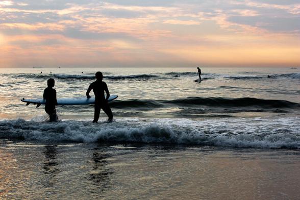 In the center left, two surfers walk from the wet sand into the sea, their bodies reflected in the wet sand. The photo was taken at sunset, so it's backlit. This makes the surfers' figures black. In the distance, on the waves toward the horizon, is another surfer. The sea extends two-quarters of the way across the frame, and above it, an orange and blue sky.