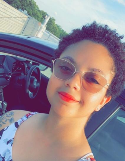A young woman with short curly black hair is smiling for a selfie while wearing a floral dress. She is standing in front of a parked car with a fence and trees in the background.