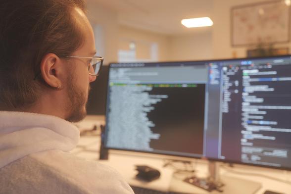 David im Ops One Büro an seinem Arbeitsplatz, im Hintergrund Bildschirme mit verschwommenem Text.