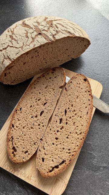 Zwei Scheiben dunkles Brot liegen auf einem Frühstücksbrettchen, links oben daneben liegt der angeschnittene Laib. Die Kruste ist bemehlt und hat ein rissiges Muster. Die Krume der Scheiben ist gleichmäßig fein mit wenigen mittelgroßen Bläschen.