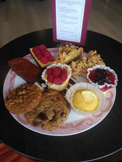 Auf einem dunkelbraunen Holztisch steht eine große, runde Kuchenplatte mit rosafarbenen Rand, auf dem sich neun Köstlichkeiten befinden (im Uhrzeigersinn):
Birnentarte mit Mandel-Pistazien-Crème (mit Mandelblättchen bestreut), Apfel-Karamell-Tarte mit Haferflocken-Zimt-Streuseln, Cheesecake mit Zwetschgenkompott obenauf, Zitronencrèmetörtchen (mit einem kleinen Stück einer Zitronenscheibe dekoriert), zwei Kekse (links, der kleinere, mit Haferflocken, Ahornsirup und getrockneten Cranberries, rechts mit Edelbitterschokolade), zwei Brownies (einmal Schokolade pur, einmal mit Frischkäse und Himbeeren obenauf und in der Mitte ein Cheesecake ebenfalls mit Himbeeren. 
Im Hintergrund ist ein Teil der Getränkekarte zu sehen.
