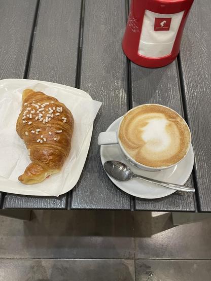A croissant, coffee, and napkin holder are on a gray table.