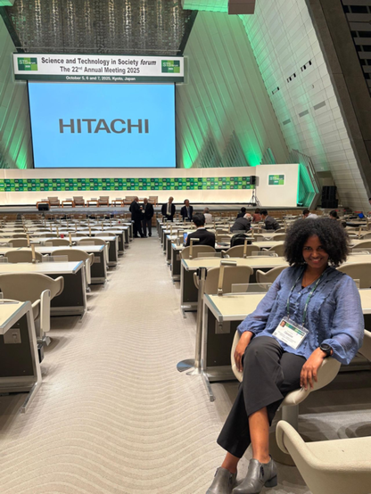 Genet sitting in the large lecture hall of STS 2025, smiling to the camera. In the bachground you see the stage and the large presentation screen.