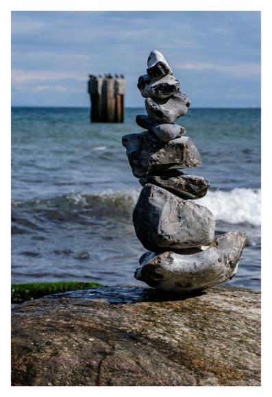 Foto im Hochformat. Im Vordergrund ist die Oberfläche eines Felsen, darauf sind acht, nach oben immer kleiner werdende, Steine gestapelt. Dahinter ist das Meer, eine Welle kommt gerade angerollt. Im Meer, Richtung Horizont, ist eine Plattform im Meer, darauf sitzen Vögel. Alles hinter dem Turm aus Steinen ist leicht unscharf.