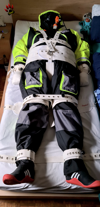 person wearing a yellow/grey/black floating suit, S10 gasmask and adidas sailing boots lying on a bed, tied in a segufix setup, full view