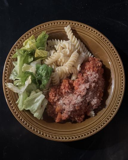 Ein brauner Teller mit Fusilli-Nudeln, Blattsalat und Bolognese, getoppt mit Hefeflocken.