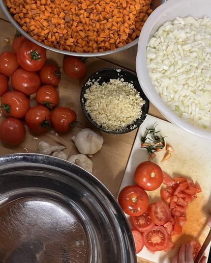 Drei Schalen voll mit geschnittenem Gemüse (Karotten, Zwiebeln, Knoblauch) und ein Schneidebrett mit Tomaten, die gerade geschnitten werden.