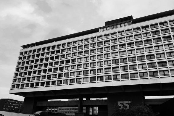 A long white modern building with many windows. It’s supported on dark concrete pylons, several storeys high, with a large sign reading “55°”.