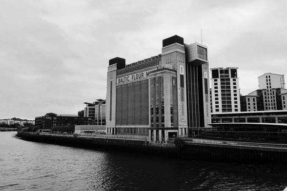 A large, square building, overlooking a river, with a sign reading “Baltic Flour Mill” in an early-20th-century style. The style of the building as a whole is pre-war industrial, and signs on the side advertise art and a restaurant.