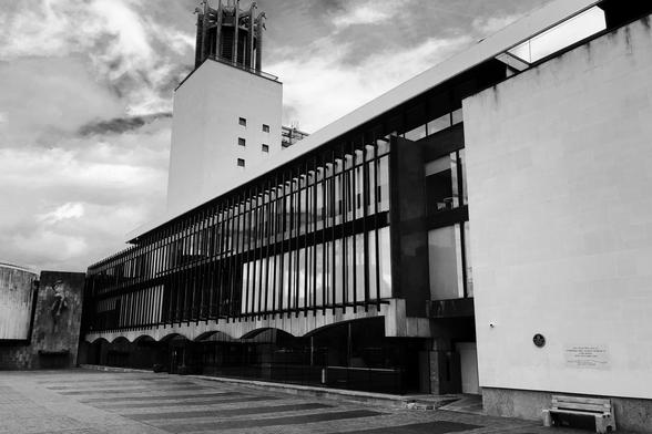 A long modernist building with glass and concrete sides. The ground floor has long concrete arches; the upper floors have windows with decorative metalwork. At the far end is a tower: a sort of modernist interpretation of a bell tower. To the left of the tower, the building starts to extend out in an L-shape, and on this part of the building is a fountain with a statue of a bearded mythological figure, larger than human height.
