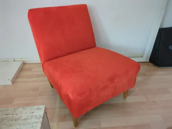 Der rotorange Sessel aus den 70er Jahren steht vor einer weißen Wand. 
Er hat Holzbeine, keine Armlehnen und sitzt sich sehr bequem.
