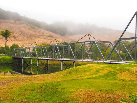 A long, metal truss bridge spans a small body of water, with a grassy hill leading up to it in the foreground. The bridge is constructed of dark metal beams arranged in a repeating triangular pattern, supported by several pillars extending into the water. The surrounding landscape features a large, sloping grassy hill covered in dry, golden vegetation, rising towards a hazy, muted background. A single tree is visible to the left of the bridge. The overall lighting is soft and diffused, with a sense of mist or fog obscuring the distant details of the landscape.