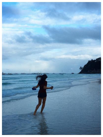 Photo of a beach, looking out to sea. Early morning sun washes the scene in blue light. A young woman in the foreground facing away from camera, her hair blown back by the breeze, holds her phone and flip-flops in her hands as she walks in ankle-deep water along the shore. The waves roll in gently along the shallow slope of the beach. At the horizon are a line of fishing boats and the shadowy mass of a tree-covered cliff rising up at right frame. The sky is overcast, with a glimpse of blue beyond gray and white clouds.