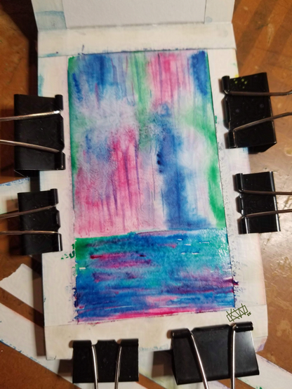 Watercolor notebook page lined with tape and held open with lots of binder clips has a very rainy, wet vibe of blurry blues, greens, & pinks.