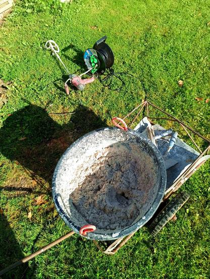 A large builders bucket (not Sir Cracksalot this time) in a self built cart. Inside is a greyish white mortar mixed not too sloppy so it can be smeared deep and not fall off a vertical wall. Some mortar tools are also on the cart. Nearby a cable drum and an electric hand mixer.