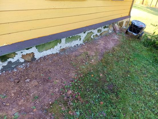 A stone foundation on the north side of a log house with yellow planks. The soil is recently disturbed from fixing the grade and the gaps between the stones are filled with fresh grayish white lime mortar. The mortar cart stands on the far corner, now less full.