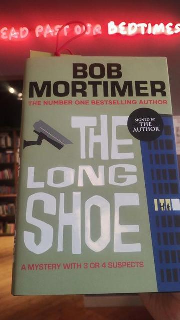 A signed hardback of The Long Shoe by Bob Mortimer, in the Edinburgh Bookshop, the store's "we read past out bedtimes" neon sign visible above and behind the book