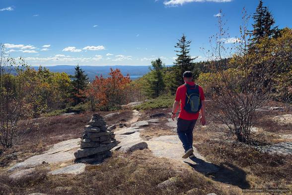 A hiker wearing a red shirt, dark pants, and a blue backpack walks along a rocky forest trail surrounded by vibrant autumn foliage. The path is uneven, scattered with stones and tree roots, and flanked by deciduous trees in shades of green, yellow, and orange. In the foreground, a small cairn made of stacked stones marks the trail. Sunlight filters through the canopy, casting dappled light across the scene. In the distance, forested hills or low mountains rise beneath a clear blue sky with scattered clouds, adding depth and a sense of destination to the tranquil, seasonal landscape.