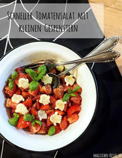 Heller Holztisch, fast völlig bedeckt von einer schwarzen Tischdecke mit weißem Spinnennetzmuster. In den unteren 2/3 des Bildes eine weiße Emailleschale mit schwarzem, dünnen Rand. Darin ein Tomatensalat, garniert mit kleinen Käse-Gespenstern und Baslikumblättern. Eine verzierte silberne Gabel und ein großer Löffel liegen über Kreuz im Salat.