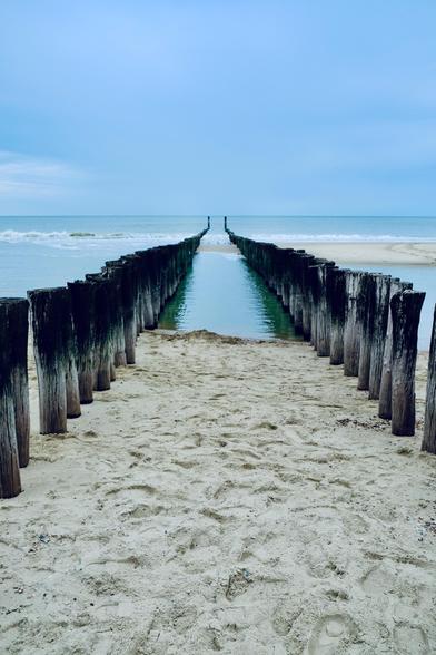Ein Blick entlang einer Reihe alter Holzpfähle, die vom Sandstrand ins Meer führen. Das Wasser ist ruhig, der Himmel leicht bewölkt und blau.