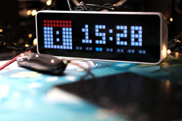 A picture of a desktop electronic clock in a pixel style. The date is on the left on an iconized calendar reading “8” and the time is 15:28. A folded pocket knife sit near the clock and a something black but too blurry to identified is on the front (it’a a PCB). The whole is on some colourful deskmat in hue of light blue and green. The background has some little LED blurry lights