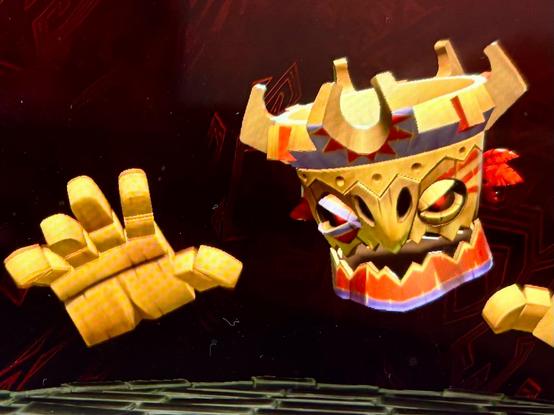 A floating, animated tiki-style mask with glowing red eyes and two large yellow hands appears against a dark red patterned background. The mask has tribal designs, red feathers on each side, and an open mouth with visible teeth. The hands are raised as if ready to attack.