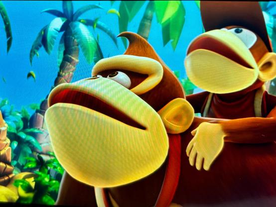 Two cartoon apes, Donkey Kong and Diddy Kong, are standing in a lush jungle environment. Donkey Kong is in the foreground looking slightly upward, while Diddy Kong, wearing a red cap, is sitting on his back with one hand resting on his shoulder. Tall green palm trees and bright blue sky are visible in the background.