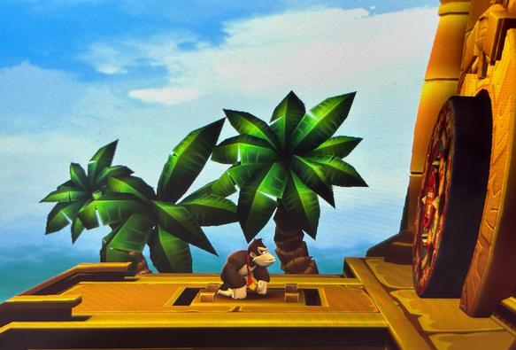 Donkey Kong is standing alone on a golden stone platform in front of a large, circular temple door. The architecture is ancient and stylized, with intricate patterns on the door. Behind him are tall palm trees and a partly cloudy sky. The scene is peaceful and quiet.