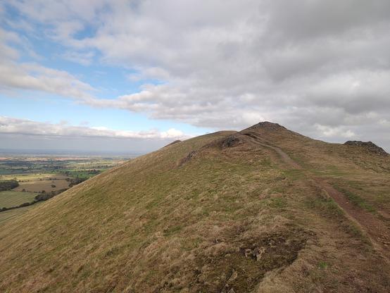 On top of a steep sided Shropshire hill with multiple peaks which stretch away into the distance. The hilltop is covered in close cropped grass and dotted with rocky outcrops and the odd sheep hearts and there. The sky overhead is cloudy but bright with some blue peeking through. On the left can be seen the rolling hills and patchwork fields of the Shropshire countryside.