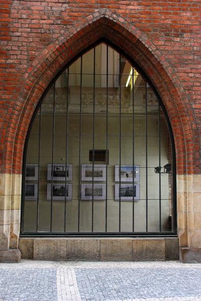 One large exterior gothic window of an art gallery. The exterior wall is marble or stone for the water table and red brick for the rest of the wall. The window has closely spaced vertical rectangular panels. Inside, we can see photos hanging on an ivory colored wall and an upper floor above. The sidewalk in front of the window is square paving stones.