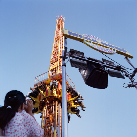 A woman looks up a photographs her friend as he ascends the yellow and orange tower of The Mega Drop.